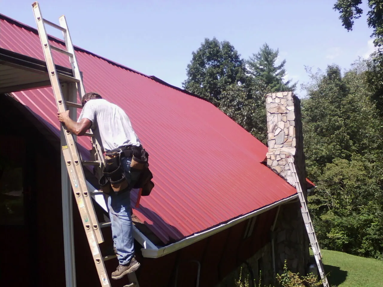 Roofer on a ladder beside a red metal roof and stone chimney — Soffit & Fascia Repair by Roofing Brothers in Foss